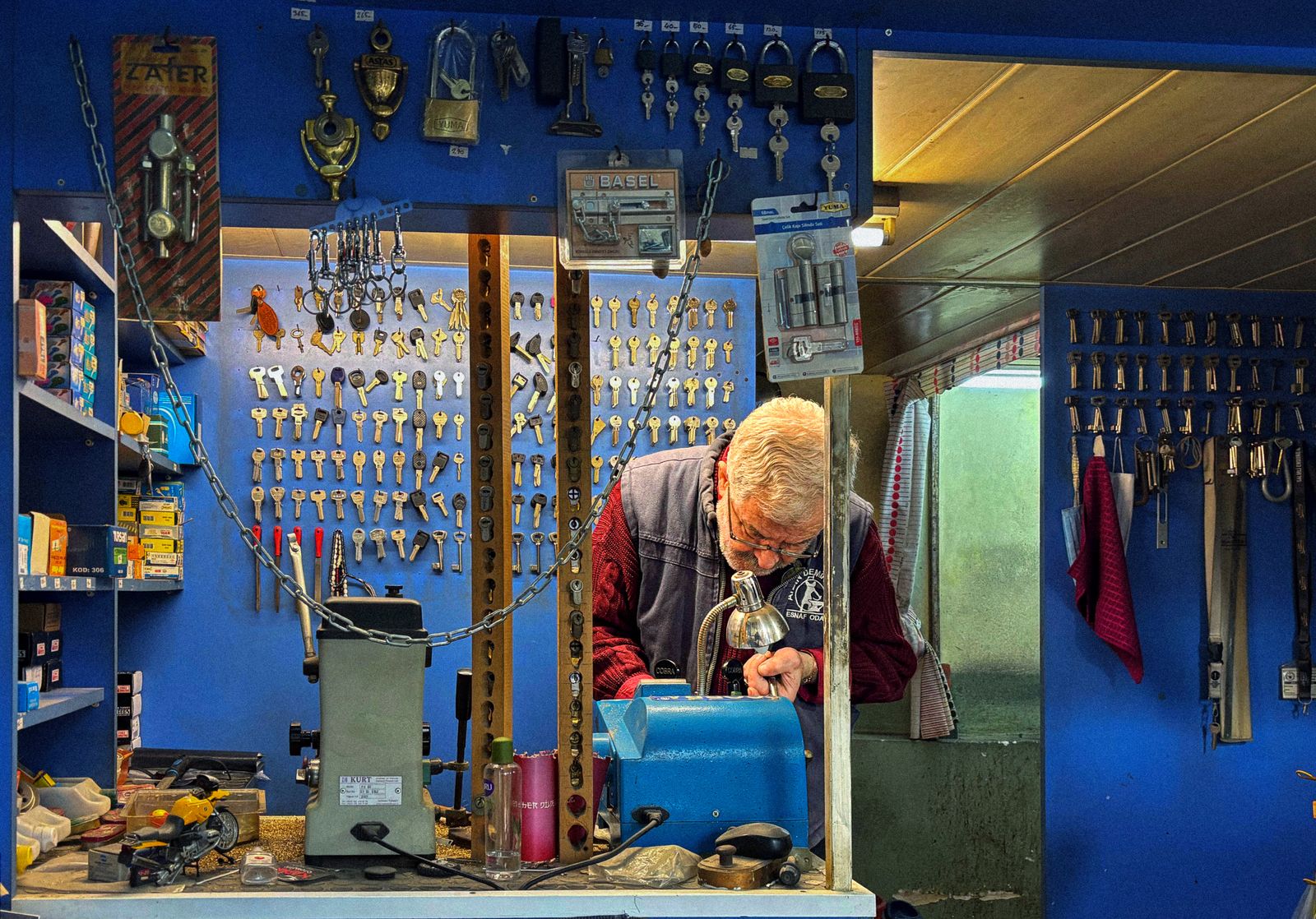 An older locksmith working at his bench in a shop full of keys and locks